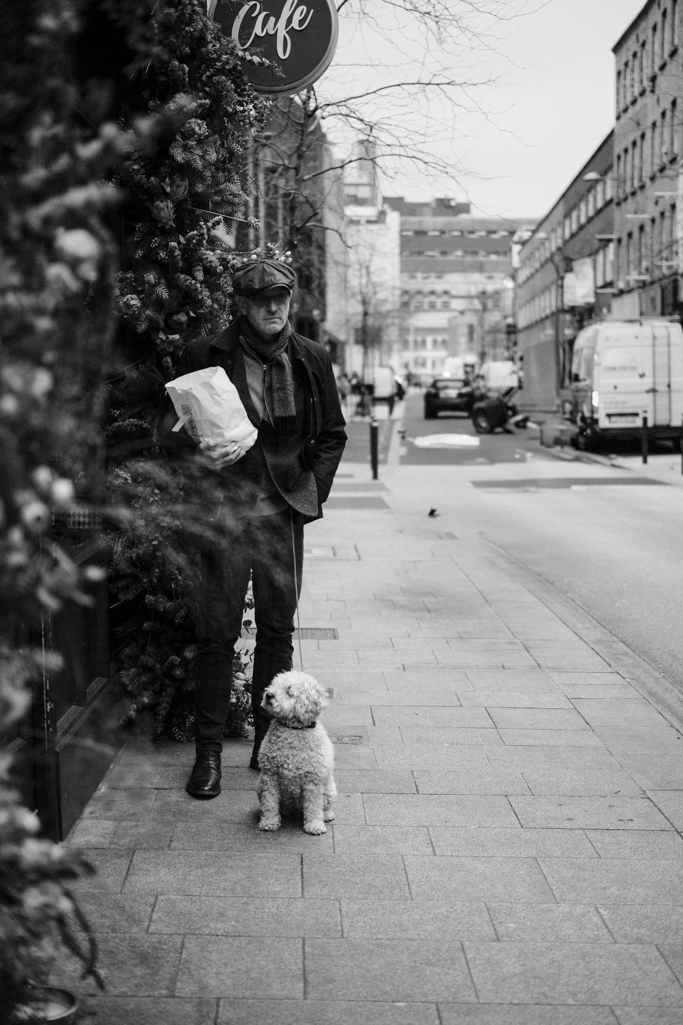 A man in a flat cap, scarf and overcoat standing on a Dublin street with a small dog