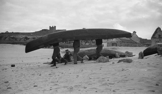Four men carrying a curragh on their shoulders across the sand on Inis Oírr, with O'Brien's Castle on the ridge behind