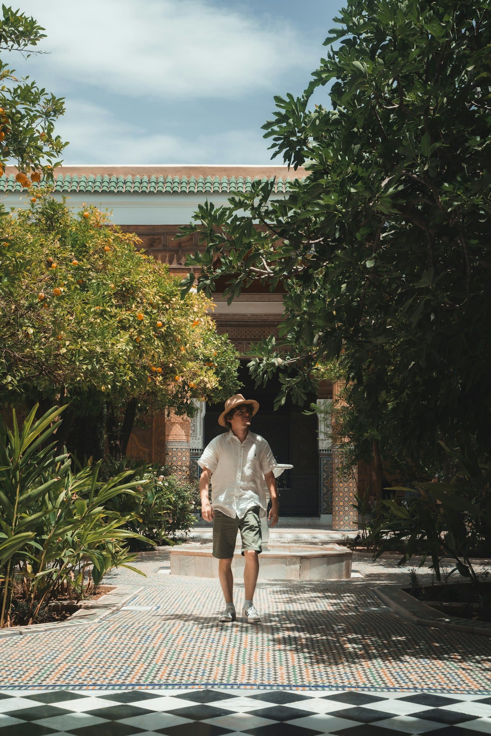 A man in a white short sleeved shirt, patterned shorts and a straw hat walks through a shaded courtyard garden