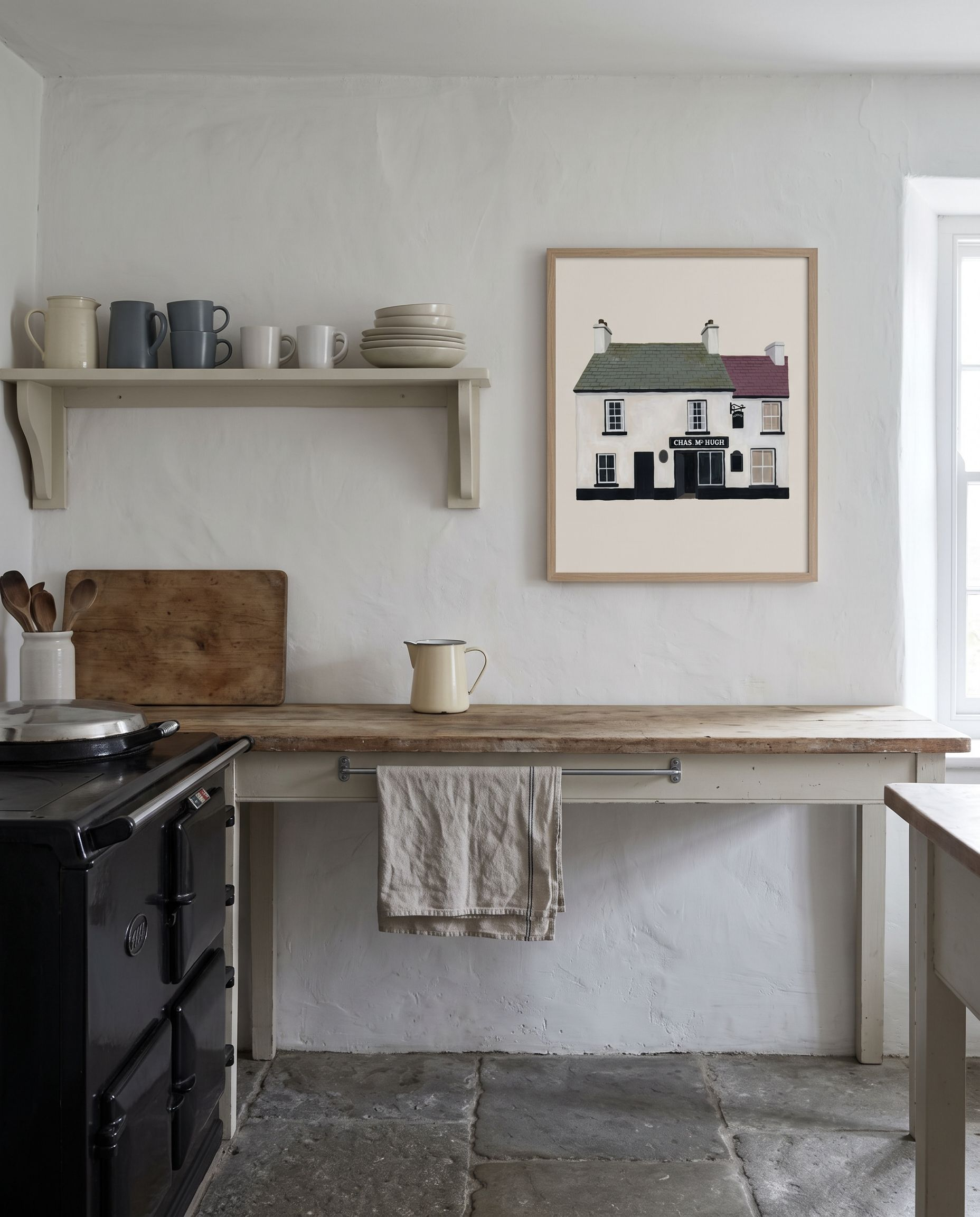 The Nancy's, Ardara print framed in oak on a whitewashed kitchen wall above a scrubbed timber worktop, beside a black cast iron range