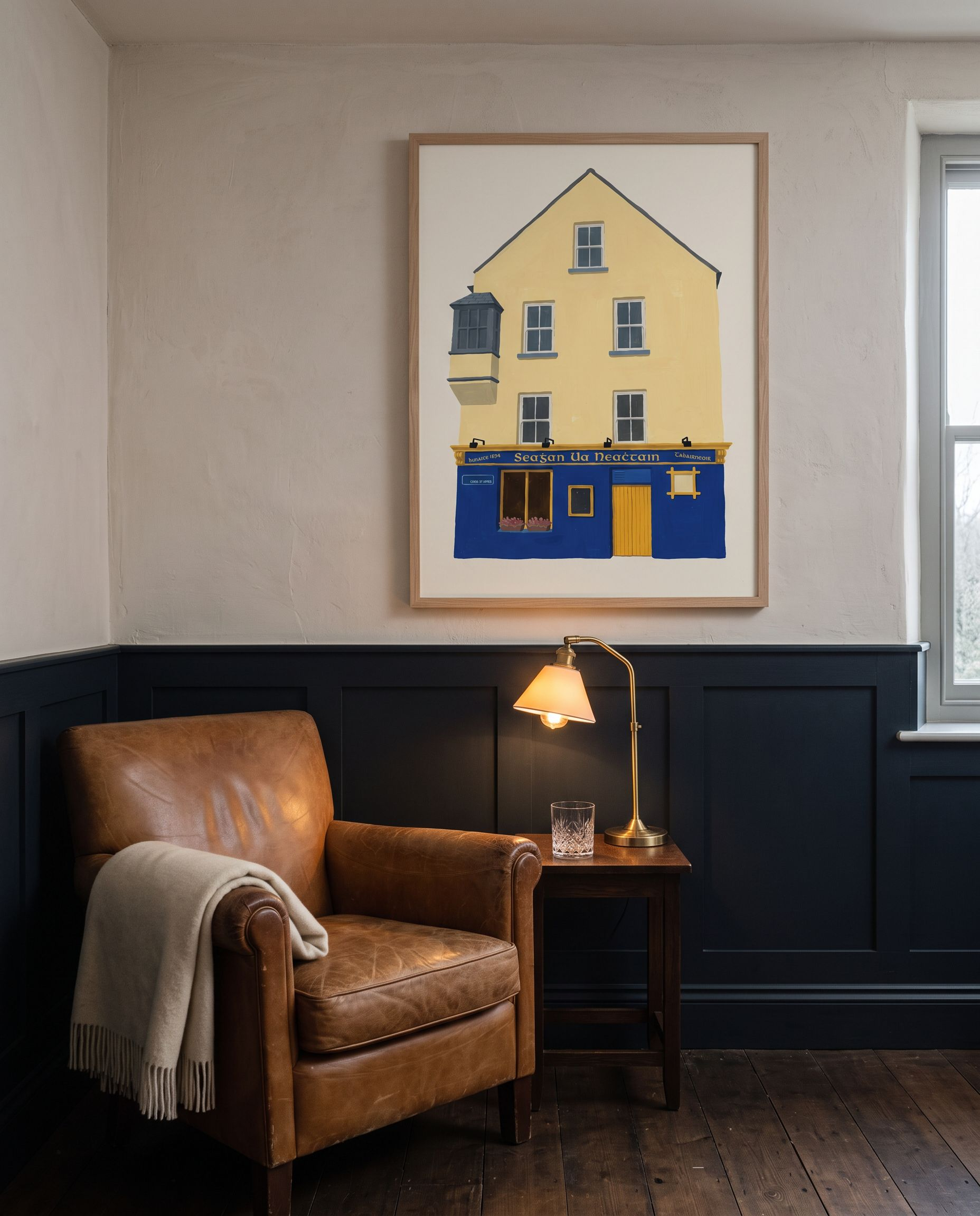 The Tigh Neachtáin print framed in oak on a plaster wall above deep blue panelling, beside a worn leather armchair and a brass lamp