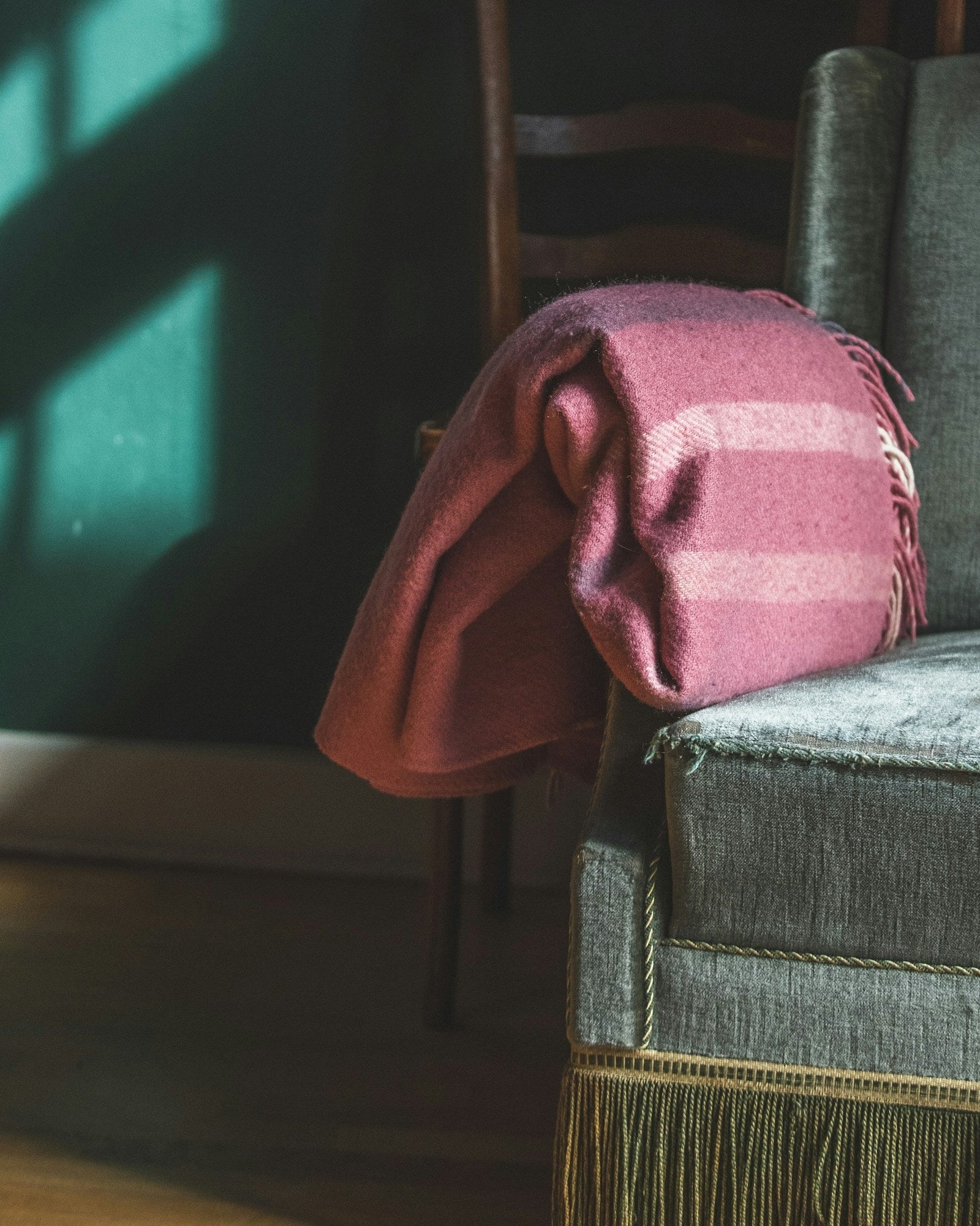 A fringed wool throw over the arm of an old armchair in a dark green room, with low window light across the wall