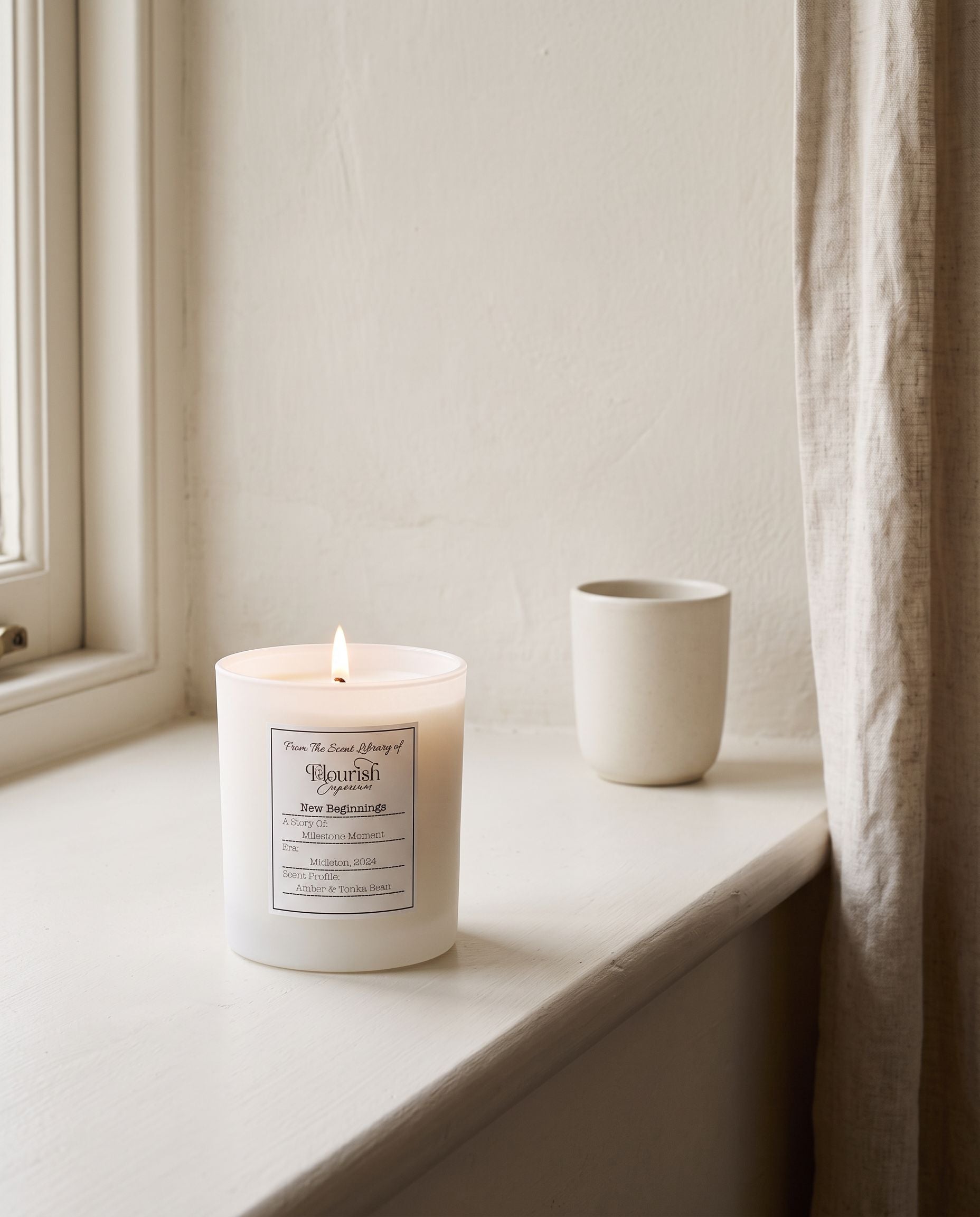 New Beginnings candle lit on a painted window sill beside a linen curtain