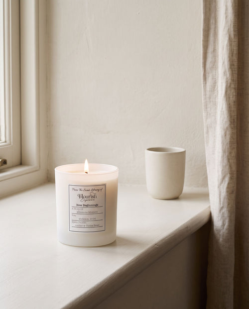 New Beginnings candle lit on a painted window sill beside a linen curtain